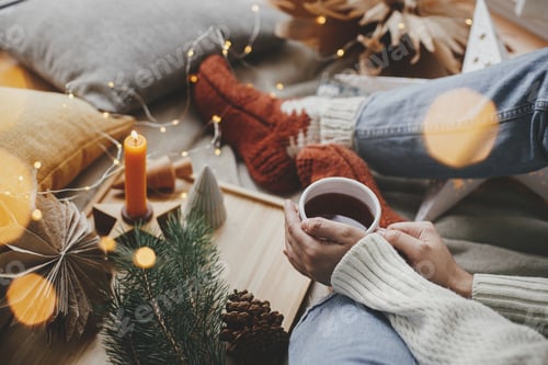 Preview: Woman hands with warm cup of tea with golden lights, christmas stars, pine, candle. Cozy hygge home