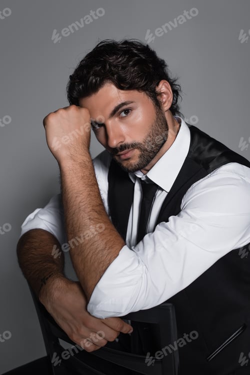 Preview: brunette man in black vest and white shirt looking at camera while sitting isolated on grey