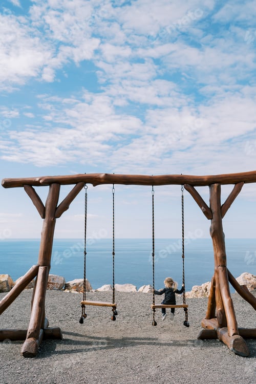 Preview: Little girl sits on a large wooden swing on a seashore and looks to the side. Back view