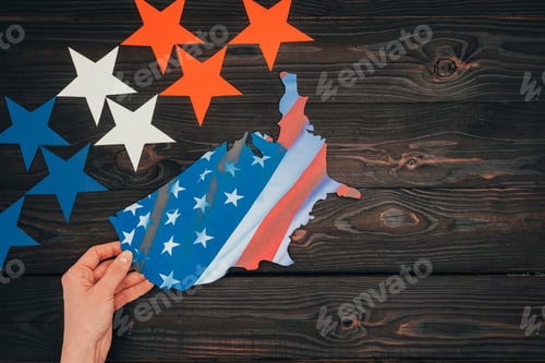 Preview: cropped shot of stars and female hand with piece of map with american flag on wooden tabletop,