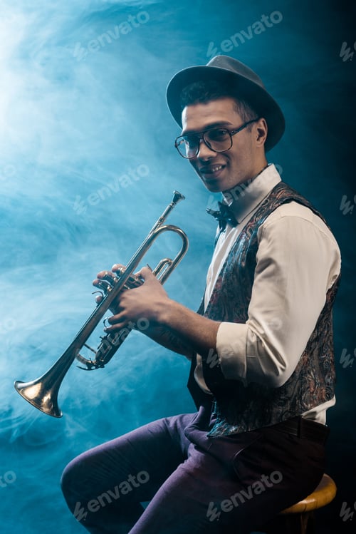Preview: happy young jazzman posing with trumpet on stage with dramatic lighting and smoke
