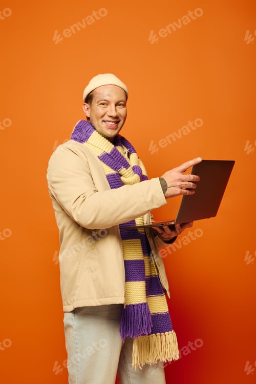 Preview: A young man with tattoos smiles while using his laptop, dressed in a unique scarf and hat