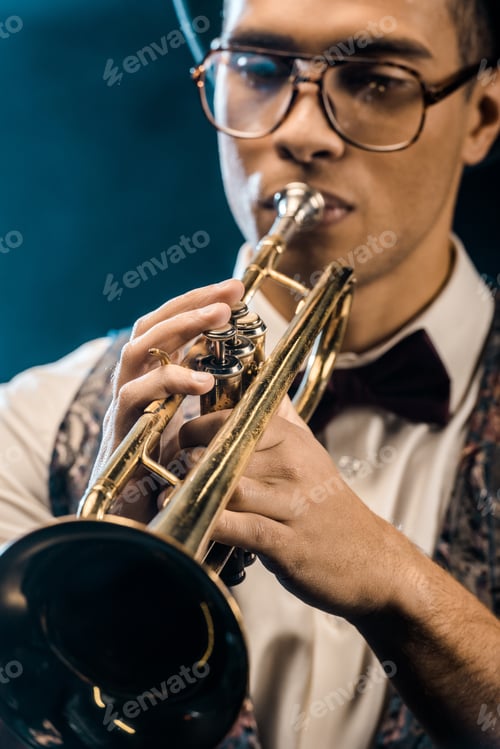 Preview: selective focus of young jazzman playing on trumpet on stage with dramatic lighting and smoke