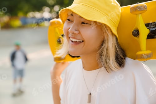 Preview: Asian girl wearing hat holding skateboard while laughing
