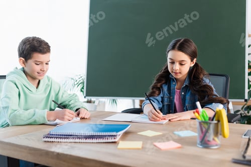 Preview: smiling boy writing in notebook near girl and chalkboard in school