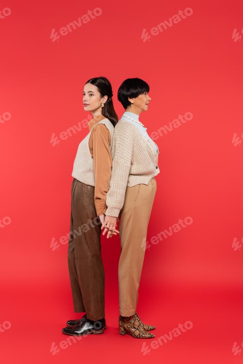Preview: full length of asian mother and daughter in stylish casual attire holding hands while standing back