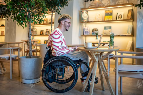 Preview: Happy pleased man on wheelchair in cafe using laptop for communication, video call, online chatting.