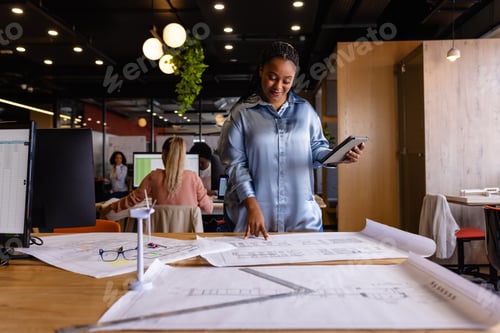 Preview: Happy plus size african american female architect using blueprints and tablet in office