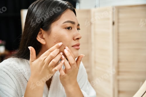 Preview: young asian woman with brunette hair examining her skin with acne in bathroom mirror, skin issue