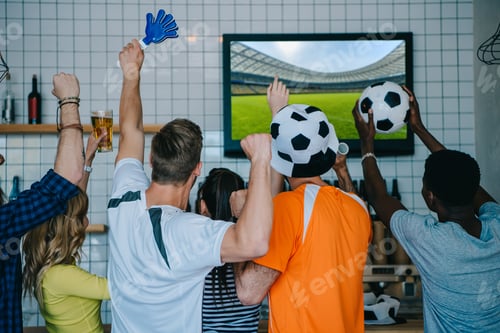 Preview: back view of football fans in soccer ball hats celebrating with hand clappers and doing yes gestures