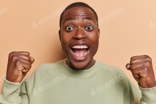Preview: Cheerful happy dark skinned man exclaims from happiness clenches fists feels like winner supports