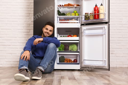 Preview: Happy young man sitting on floor near open refrigerator indoors