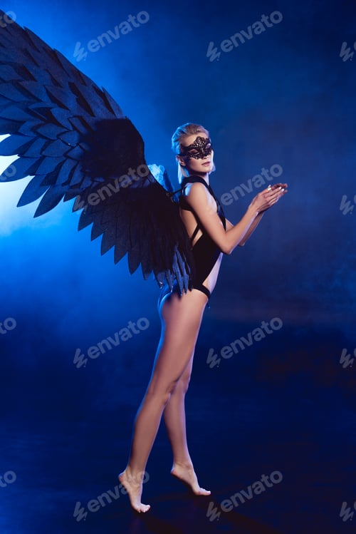 Preview: beautiful sexy woman with lace mask and black angel wings standing on tiptoe on dark blue background