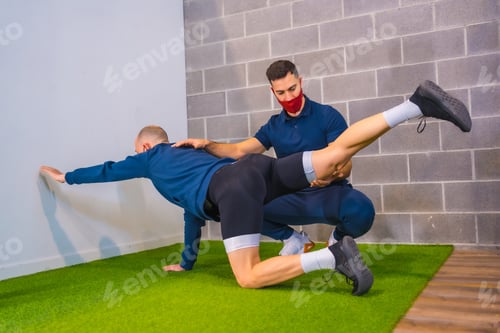 Preview: Personal trainer in gym correcting stretching of a student on the floor in the coronavirus pandemic