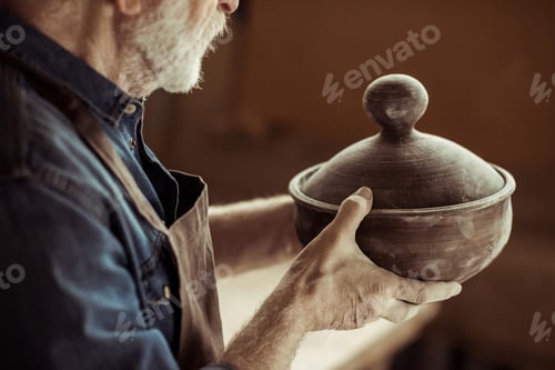 Preview: Senior potter in apron examining ceramic bowl at workshop