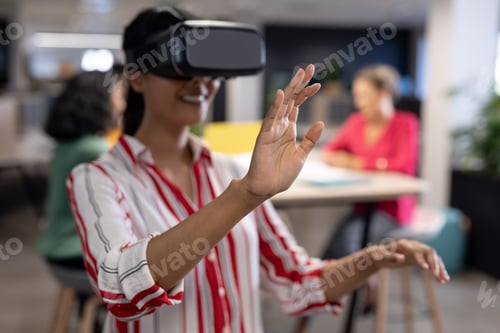 Preview: Young biracial businesswoman experiencing virtual reality simulator while gesturing at workplace