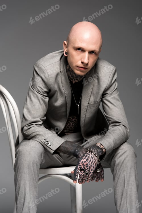 Preview: handsome confident tattooed man in suit sitting on chair and looking at camera isolated on grey