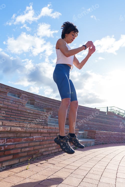 Preview: Fit mature woman jumping during outdoor exercise near brick bleachers