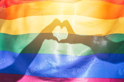 Preview: LGBTQ rainbow colors flag and two hands making a heart shape shining by sunlight