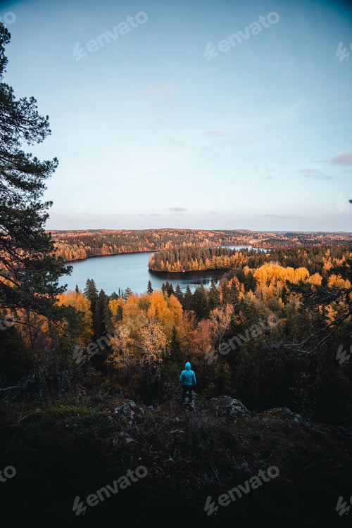 Preview: Vertical of a man standing on a mountain looking at the foliage trees and a lake