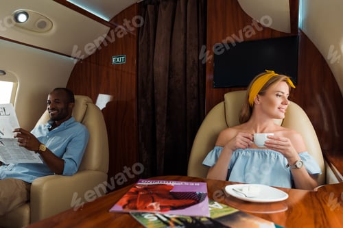 Preview: cheerful woman holding cup with drink near african american man in plane