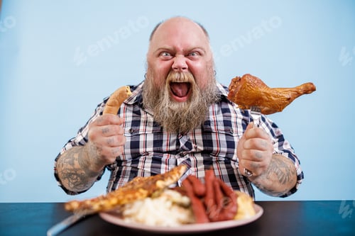 Preview: Emotional obese man holds sausage and smoked chicken leg sitting at table