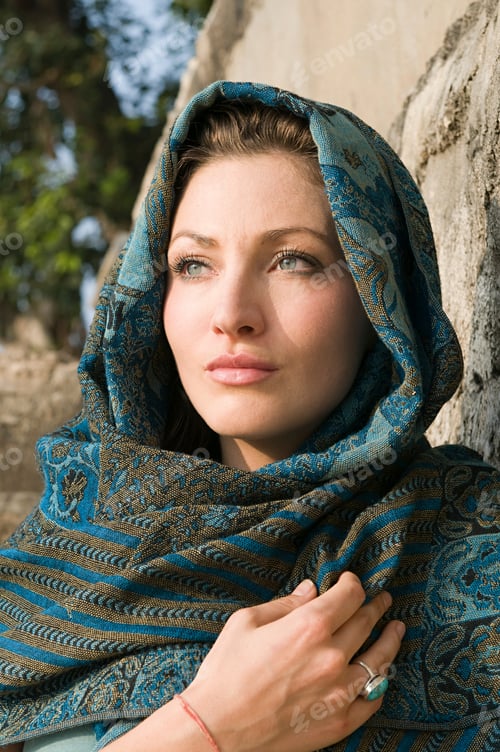 Preview: A thoughtful woman draped in a blue patterned shawl gazes into the distance.