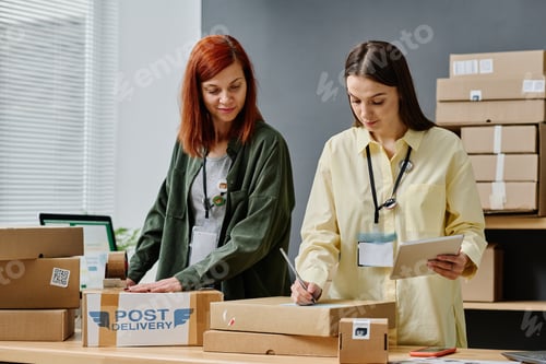 Preview: Young woman with tablet writing down address of receiver on packed box