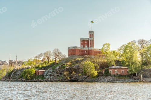Preview: Red brick Stockholm Castle with a flag tower on an island against a clear blue sky