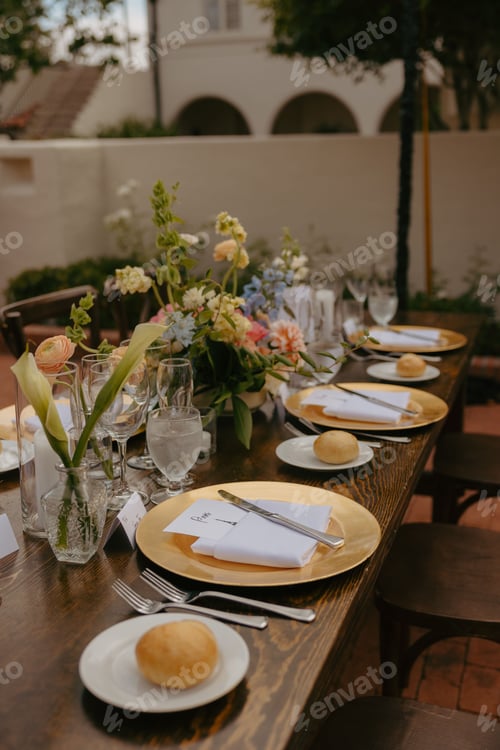 Preview: Wooden table set for a wedding reception with gold chargers, white plates, and blue napkins.