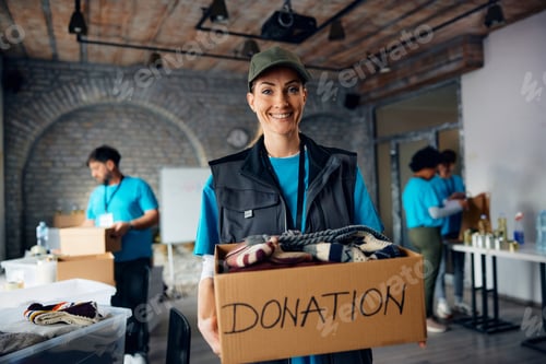 Preview: Happy female volunteer with carboard box of donated clothes at community center looking at camera.