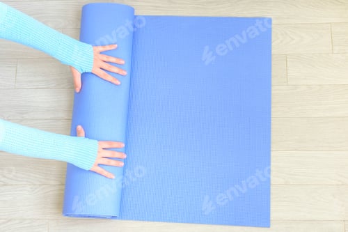 Preview: Female hands roll up rubber blue exercise mat on the floor close-up, top view.