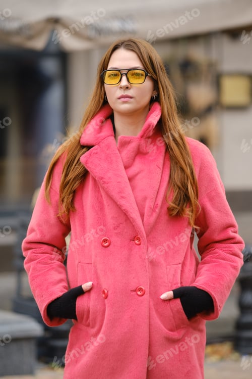 Preview: A young woman in a pink coat and yellow sunglasses stands confidently outdoors in a city setting