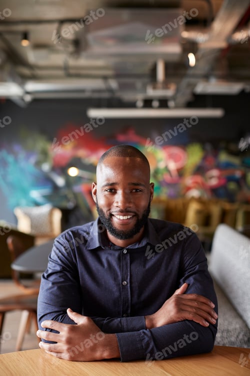 Preview: Mid adult black male creative sitting at a table with arms crossed smiling to camera, close up
