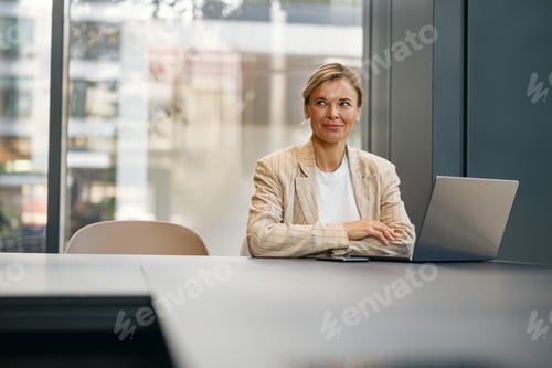 Preview: Focused business woman working on laptop sitting the desk on office background and looks camera