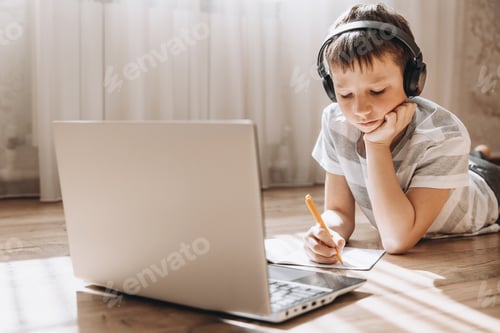 Preview: Teenage boy in wireless headphone lying on floor using laptop having online lesson at home.