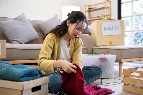 Preview: Cute Asian woman sitting next to sofa in living room sorting unwanted clothes for donation.