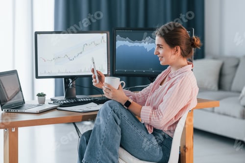 Preview: Sitting with cup of drink and smartphone in hands. Female stock broker in casual clothes is working