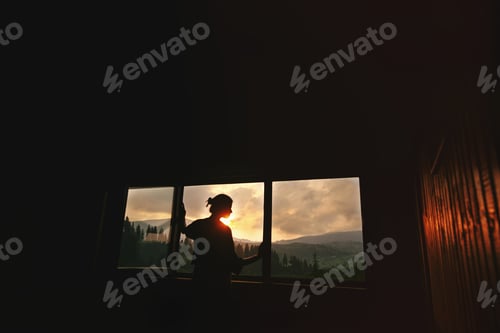 Preview: Silhouette of woman in wooden cottage window with view on sunset in mountains