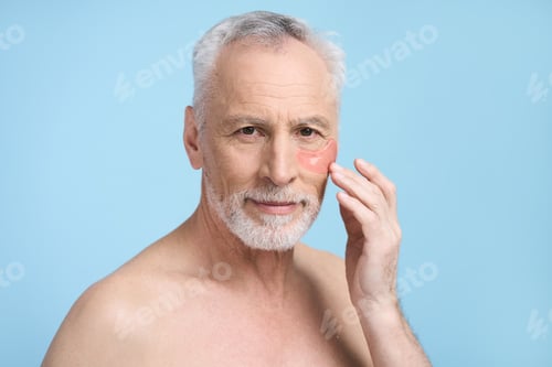 Preview: Handsome senior man applying hydrogel patches under eyes, isolated on blue background. Male beauty