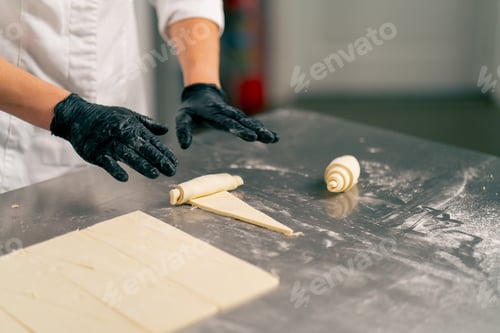 Preview: Close-up shot of a chef's hand in gloves rolling raw cut dough into croissant shapes for baking