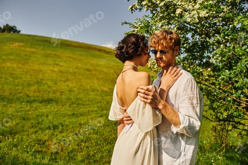Preview: boho style, happy multiethnic newlyweds in sunglasses and wedding gown hugging in countryside