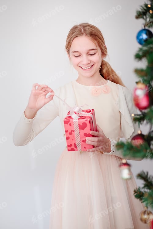 Preview: Happy smiling girl wearing pink dress standing behind a tree and unpacking Christmas gift