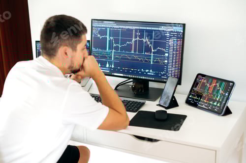 Preview: Concentrated male trader sitting at desk at home office monitoring stock market looking at monitors
