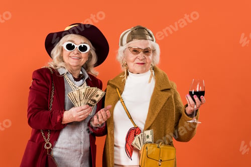 Preview: cheerful senior woman holding money near friend with glass of wine in hand isolated on orange
