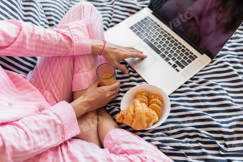 Preview: cute young pretty blond woman in pink pajamas sitting on bed working on laptop, freelancer at home