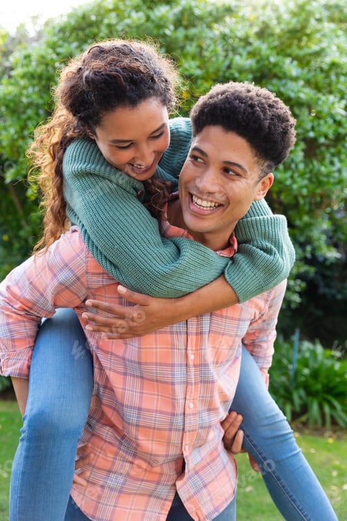 Preview: Happy biracial couple having fun piggybacking in garden and smiling