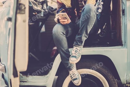 Preview: Vintage fashion style woman with sneakers sitting outside her old blue van and checking mobile
