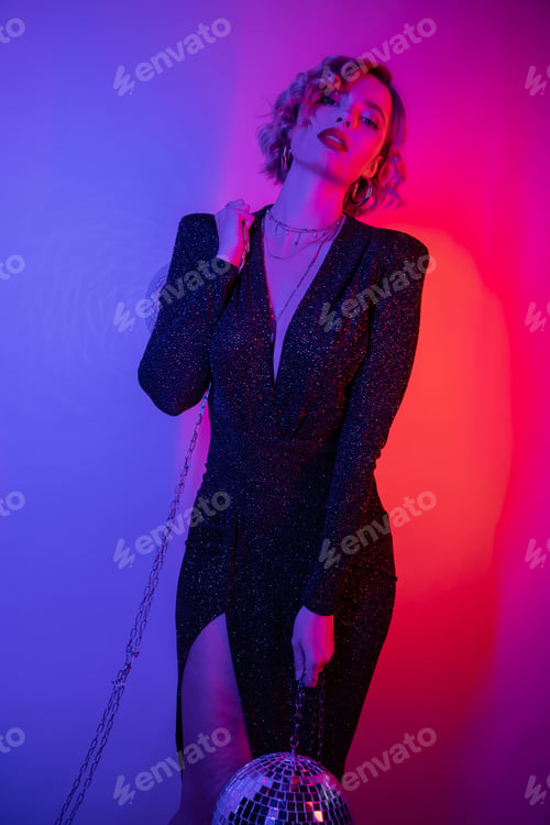 Preview: young woman with blonde hair standing in black tight dress and holding chains with disco ball on