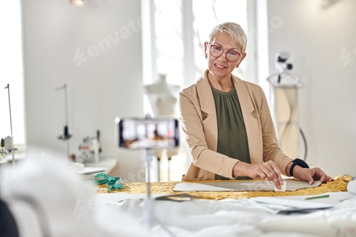 Preview: Positive middle aged woman draws pattern on fabric shooting video for blog in workshop